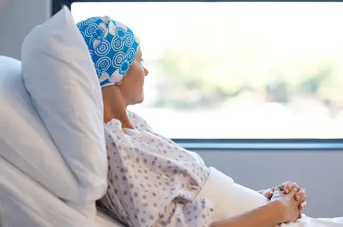 A female cancer patient lies in a hospital bed and looks out of the window. Her head is covered