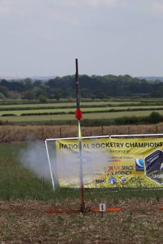 A small rocket takes flight in a field in front of a sign that sayds 'national rocketry championship'