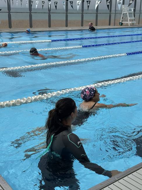 A woman in a wetsuit and woman in a swimsuit and swimming cap swim in an open water swimming pool. Some other swimmers in the background.