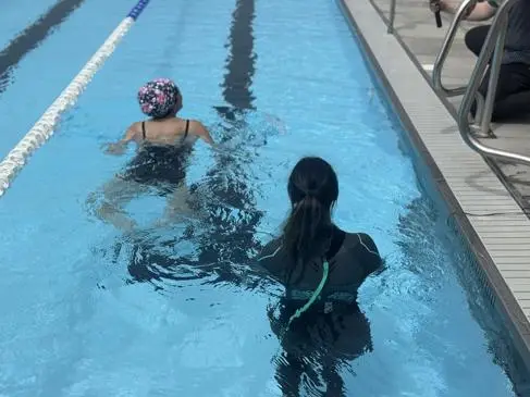 A woman in a wetsuit and woman in a swimsuit and swimming cap swim in an open water swimming pool.