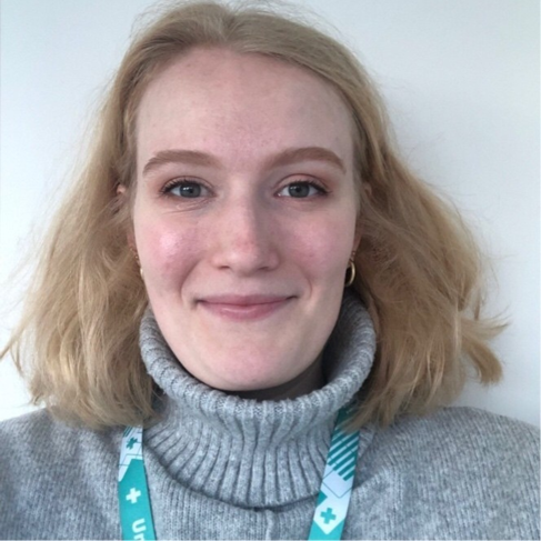 A young woman smiles. She has a blonde bob and wears a grey high-neck jumper