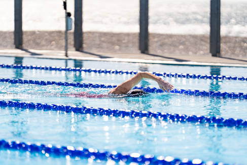 An outdoor swimming pool with lanes, with one swimmer in one swimming overarm in one of them.