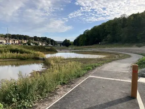 Rainscape, with a path flowing around it on the right and a lady walking on it. Houses in the distance.