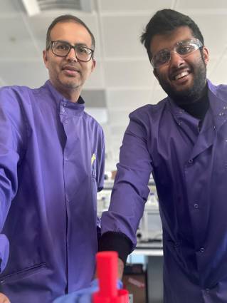 Two men wearing blue lab coats and protective glasses smiling at the camera