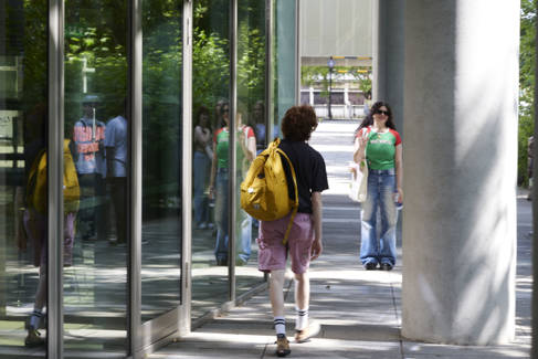 Two students walking in opposite directions between a glass window and a concrete pillar.