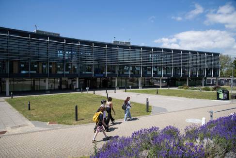 University campus on a sunny day, with a modern glass building and a green social area with students walking