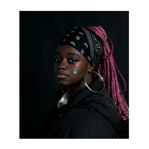 Young black woman turns to face the camera. She is wearing a large hoop earring, a black bandana and has her pink hair tied up.