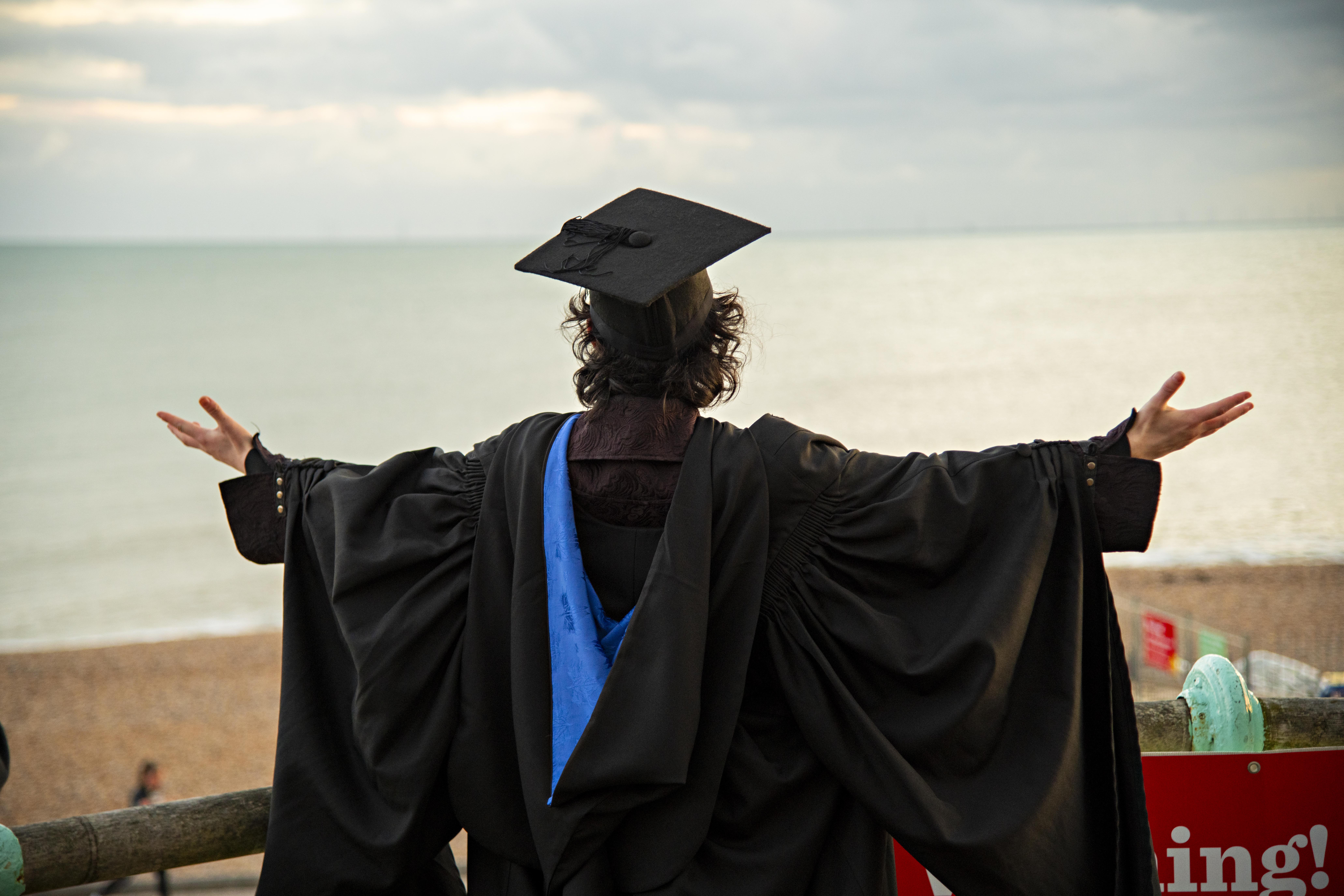 A young university graduate wearing a cap and gown faces away from the camera. His arms are extended. He stands in front of the sea.