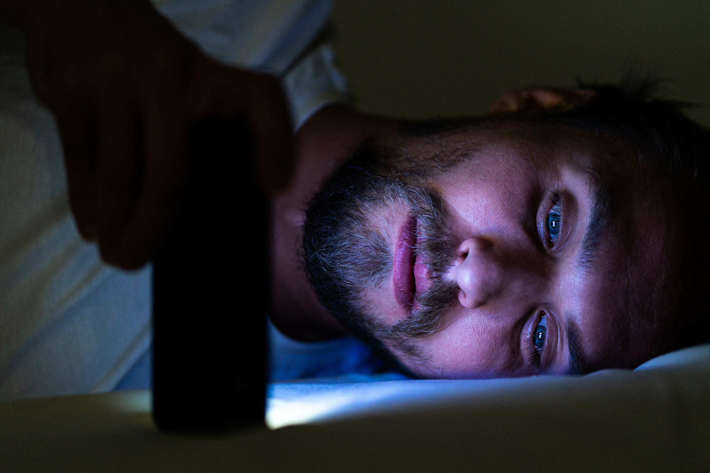 Bearded young man looks at his phone at night. Blue light from the phone is all over his face