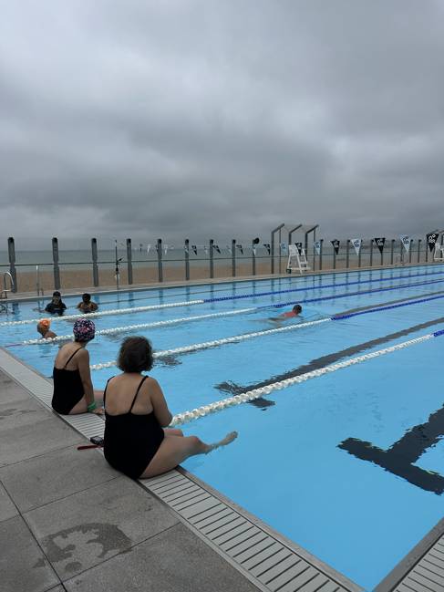 swimming people with people sat on the edge, and some swimming. The sky is grey in the background