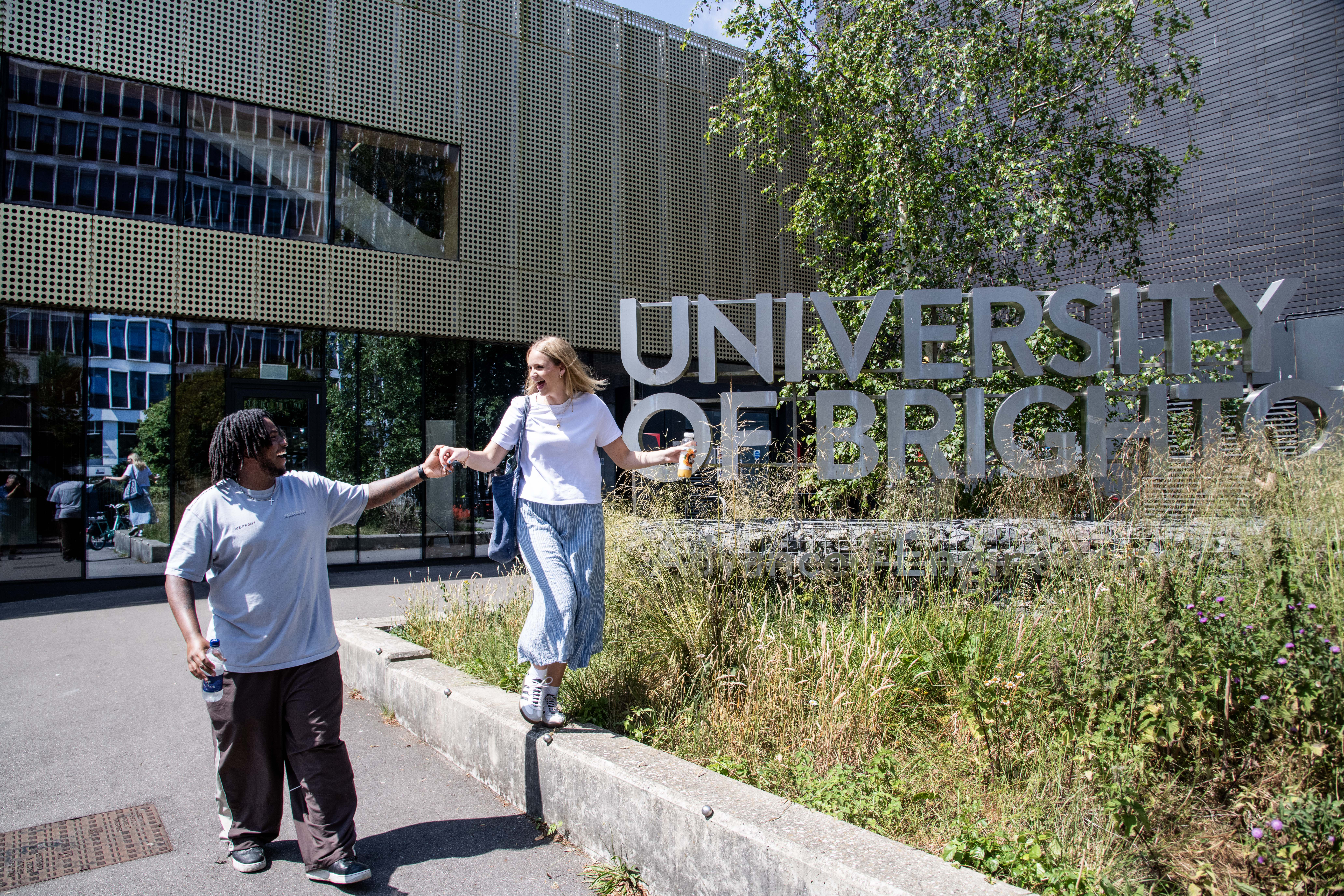 Two students holding hands in front of a modern building and sign reading 'the University of Brighton'