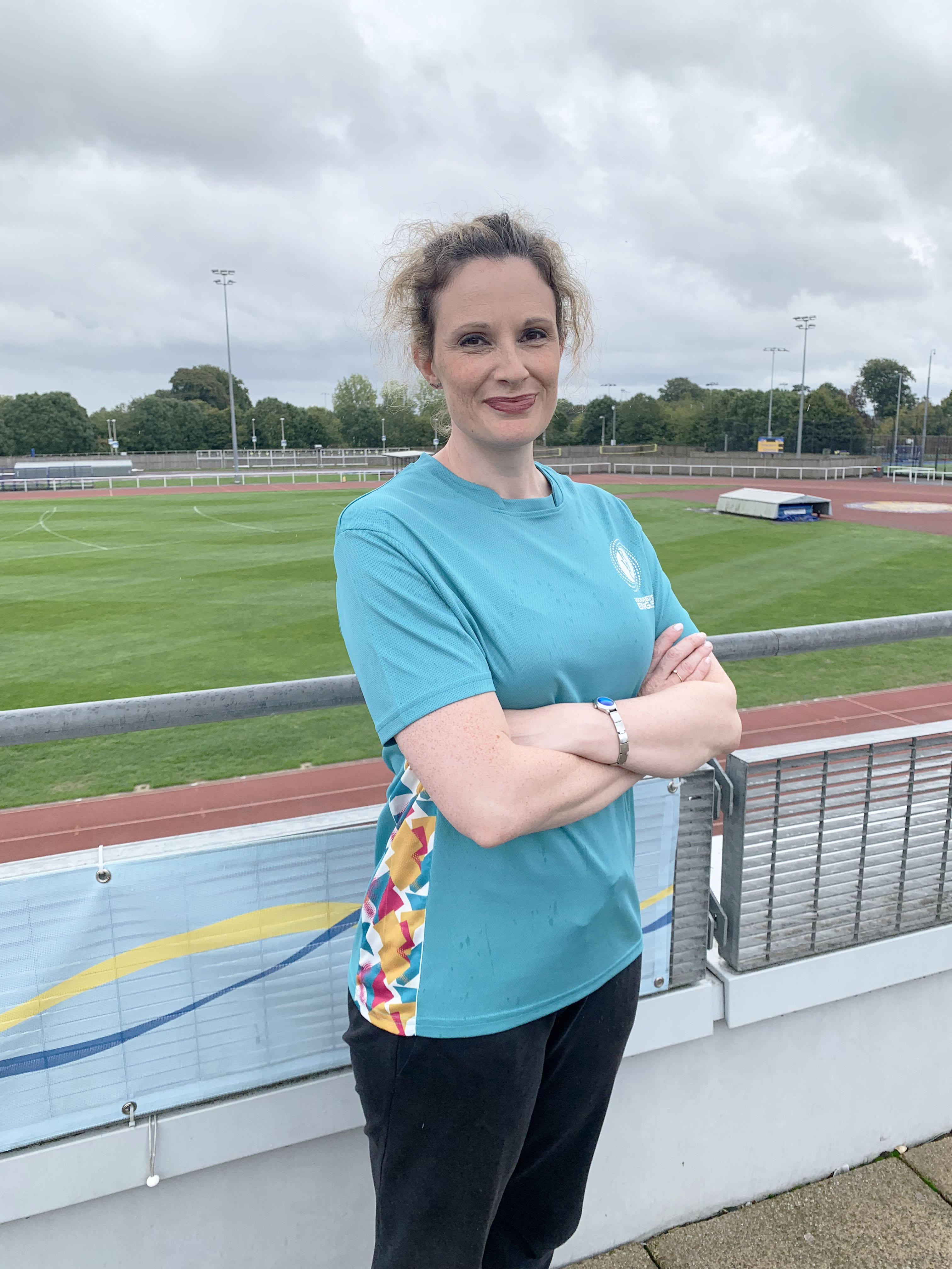 Woman wearing a blue sports top stands in front of a green sports field with her arms folded