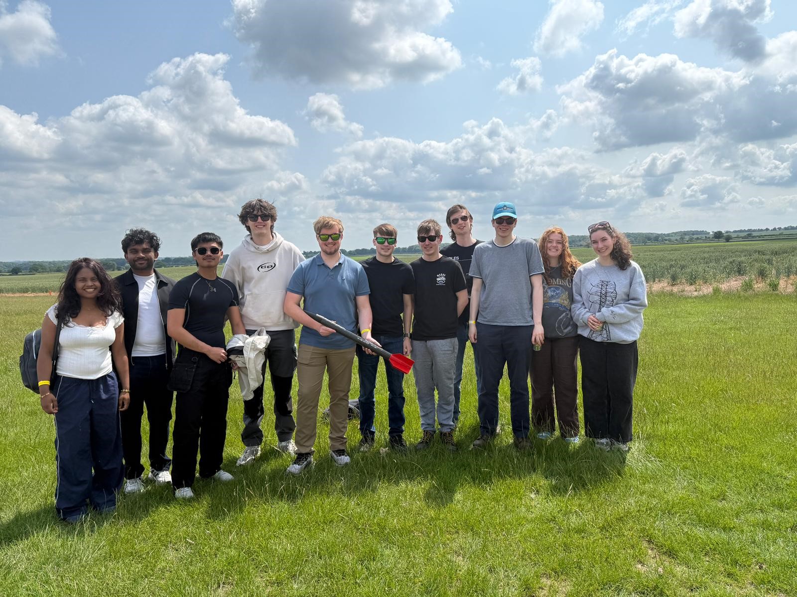Young students stand in a green field, in a row, holding a small rocket