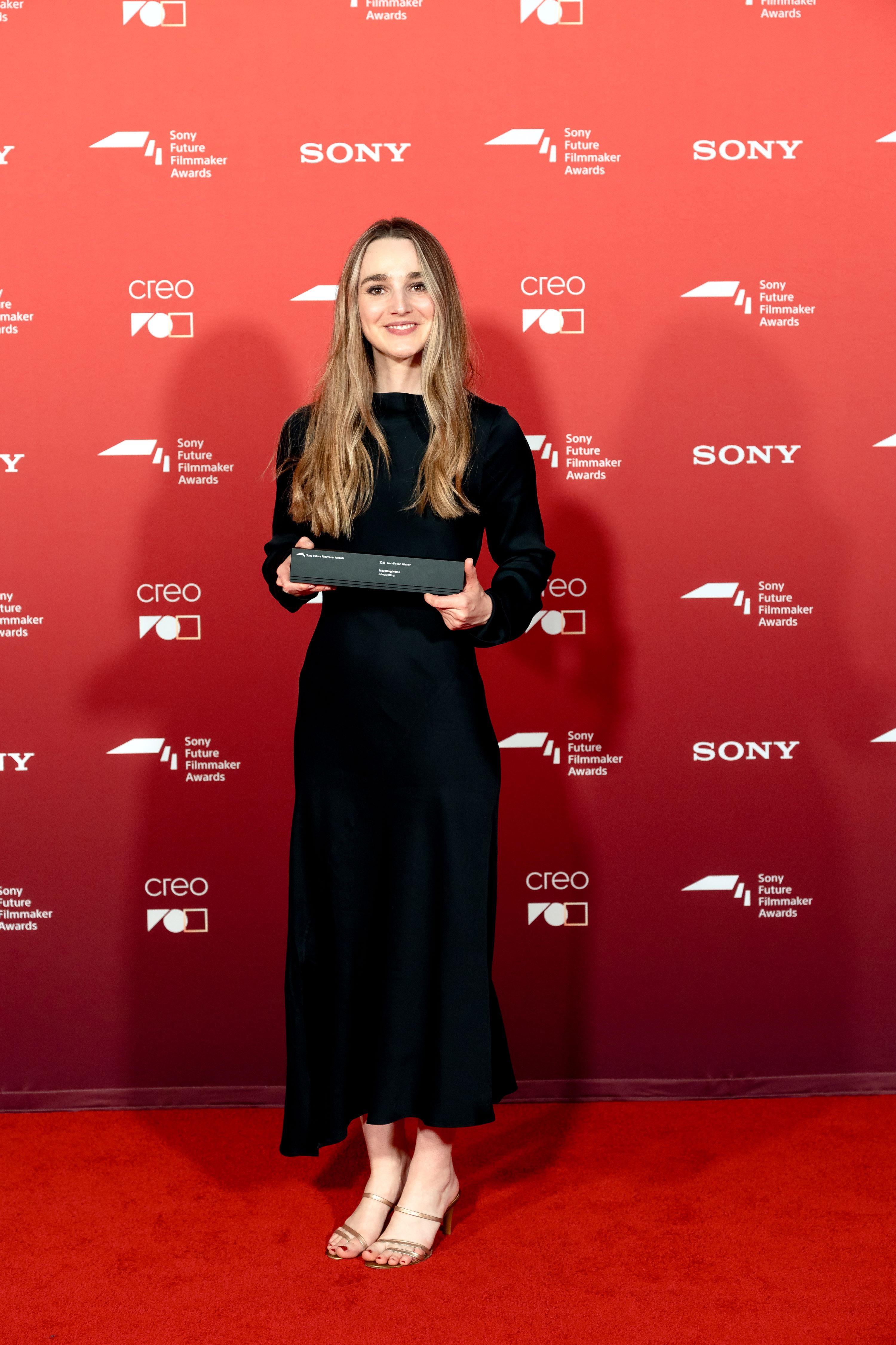 Young woman in black dress holds award in front of red media wall, on red carpet