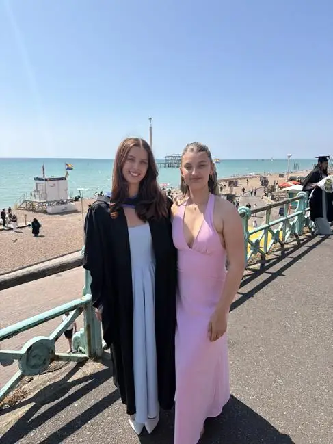 A graduate in a cap and gown stands next to her sister in a pink dress
