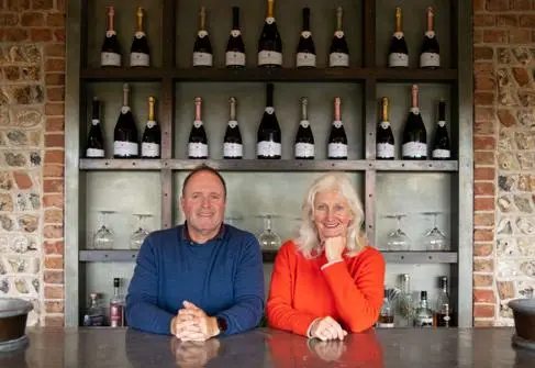 A man in a blue jumper and a woman in red jumper smile in front of a brick wall and a cabinet full of wine