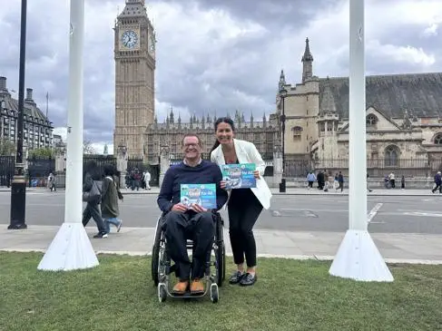 A man in a wheelchair and a woman smile outside Big Ben holding a report