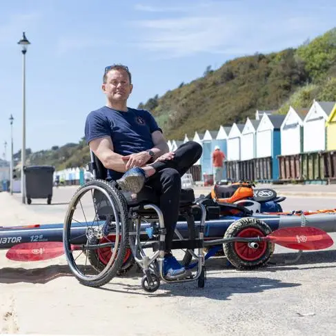 A man sits in a wheelchair on the beach, with a paddleboard in front of him