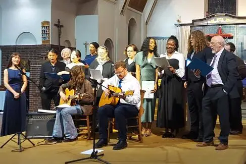 A mixed group of students and staff sing and play instruments on stage