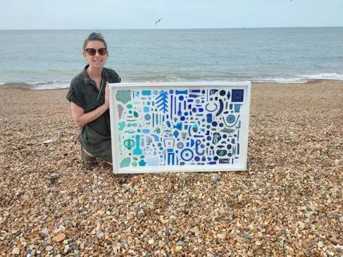 A woman holds a piece of artwork on the beach. It's made of blue, navy and black plastic