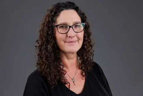 A woman with dark curly hair wears glasses and smiles at the camera