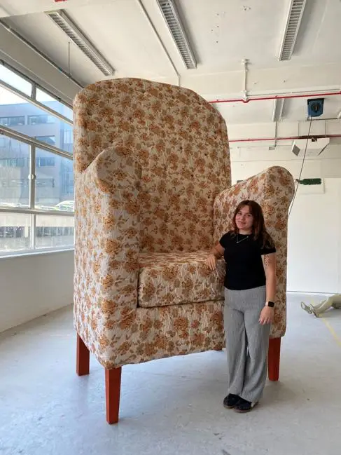 A young woman (Kayleigh Hogarth) stands next to a giant chair