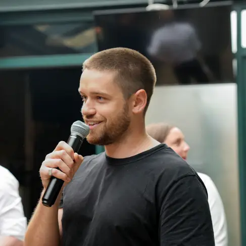 A young man with short hair and wearing a black t-shirt, holds a mic to his lips