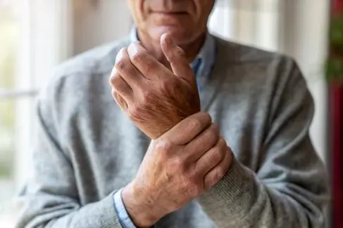 An elderly man wearing a jumper holds one wrist in his other hand. You can't see his face