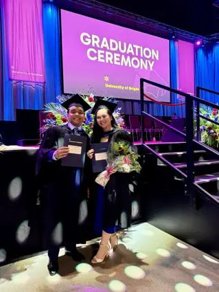 Two students smile in caps and gowns at the University of Brighton graduation ceremony. They pose in front of the stage