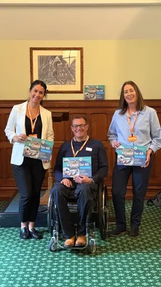 Two women and a man in a wheelchair smile holding a report called 'A Coast for All'