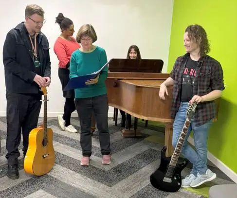 University students stand around a piano with guitars. Someone sits at the piano.