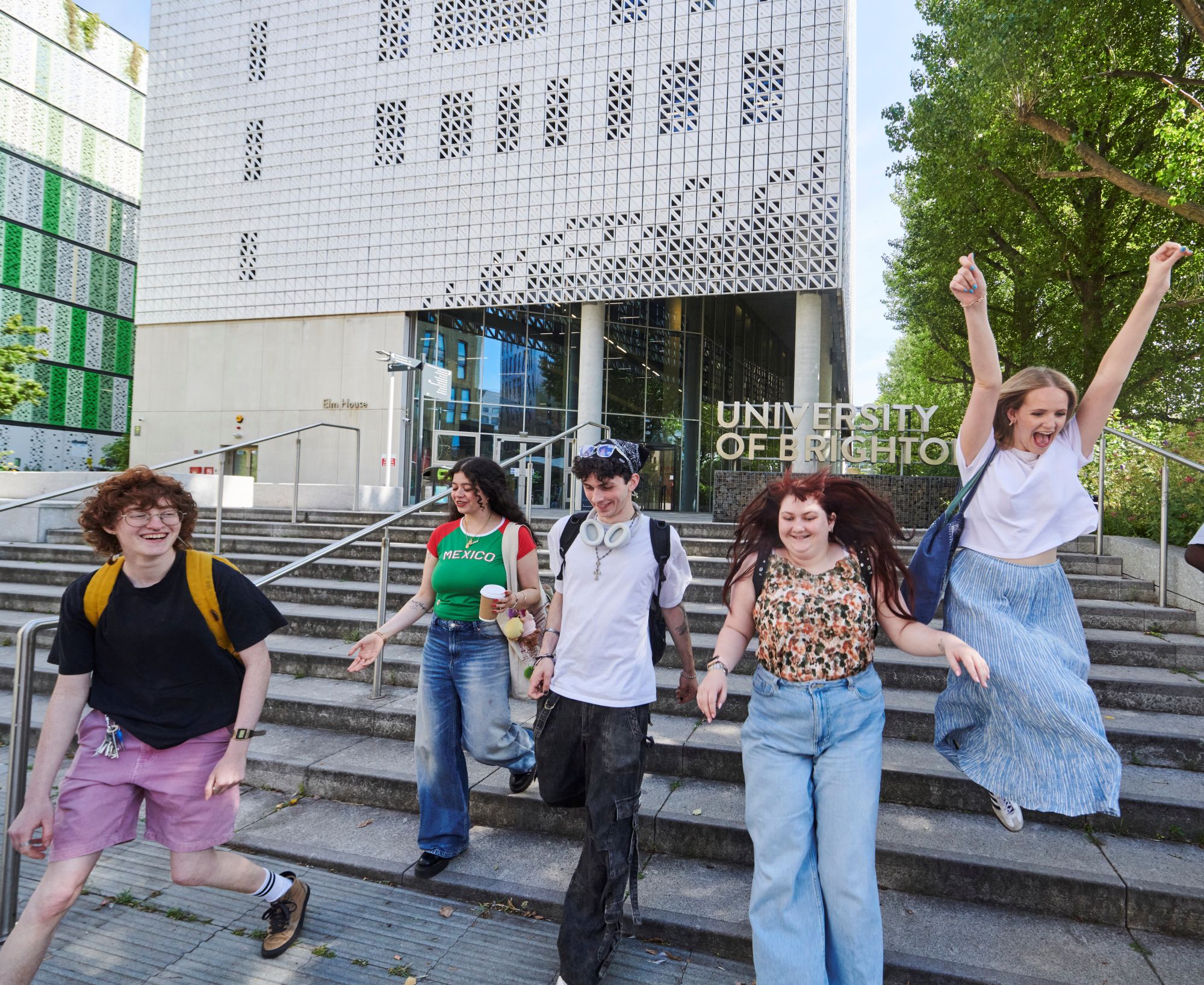 A group of students smile, walk and jump as they walk down stairs in front of a University of Brighton building and large sign