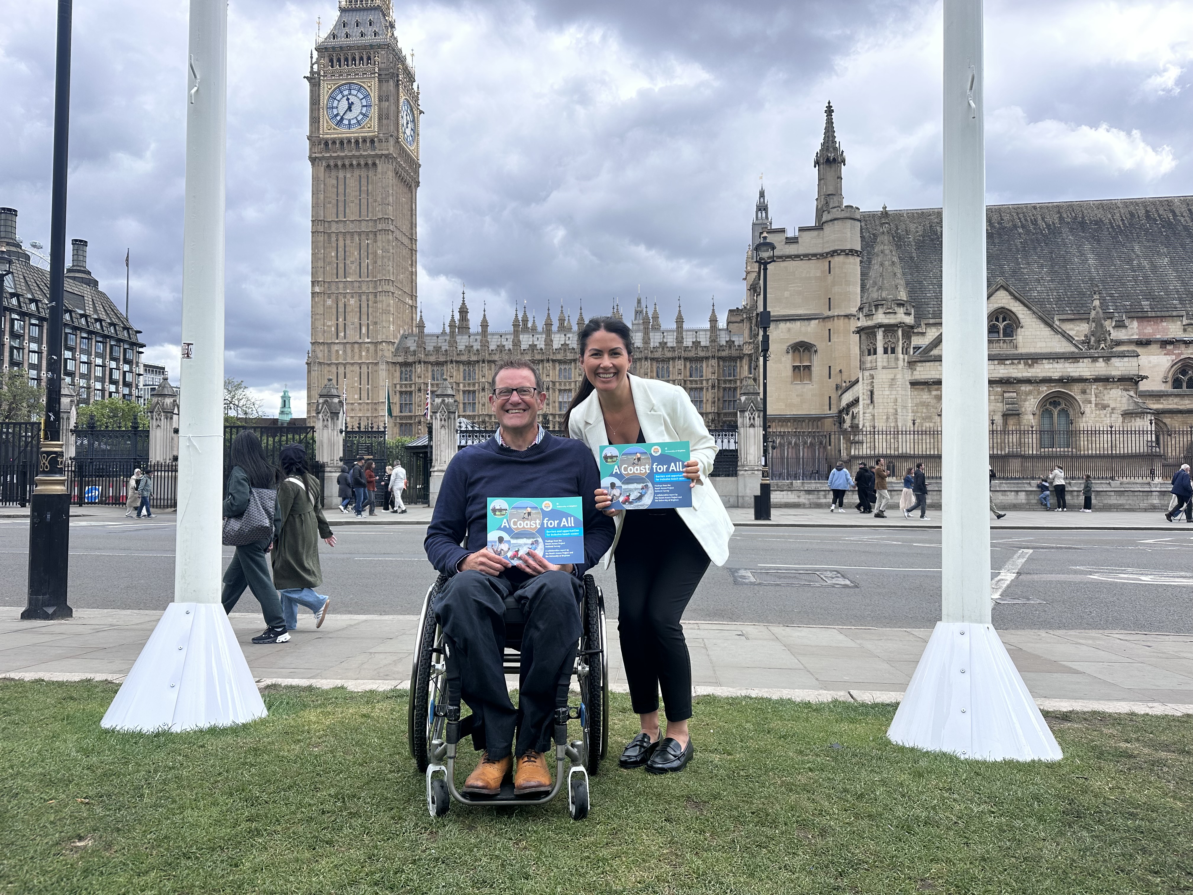 A man in a wheelchair and a woman smile outside Big Ben holding a report