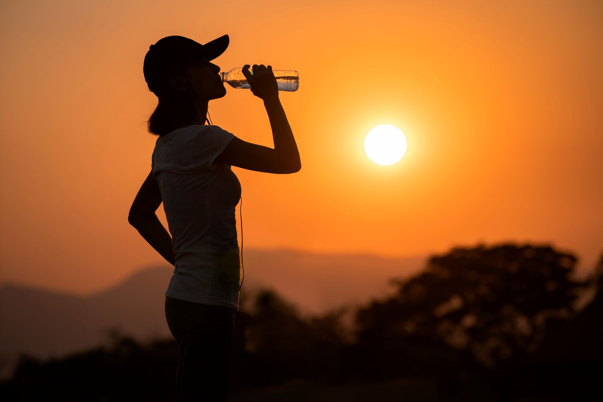 A woman drinks water with the sun glaring in the background