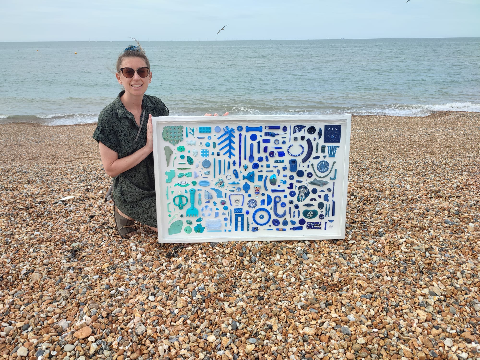 A woman holds a piece of artwork on the beach. It's made of blue, navy and black plastic