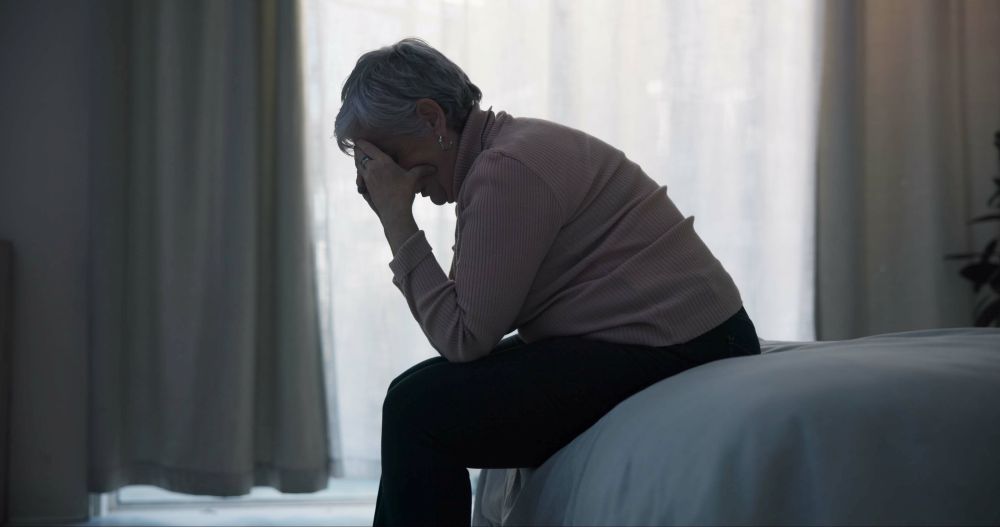 A woman holds her head in her hands as she sits on the end of a bed