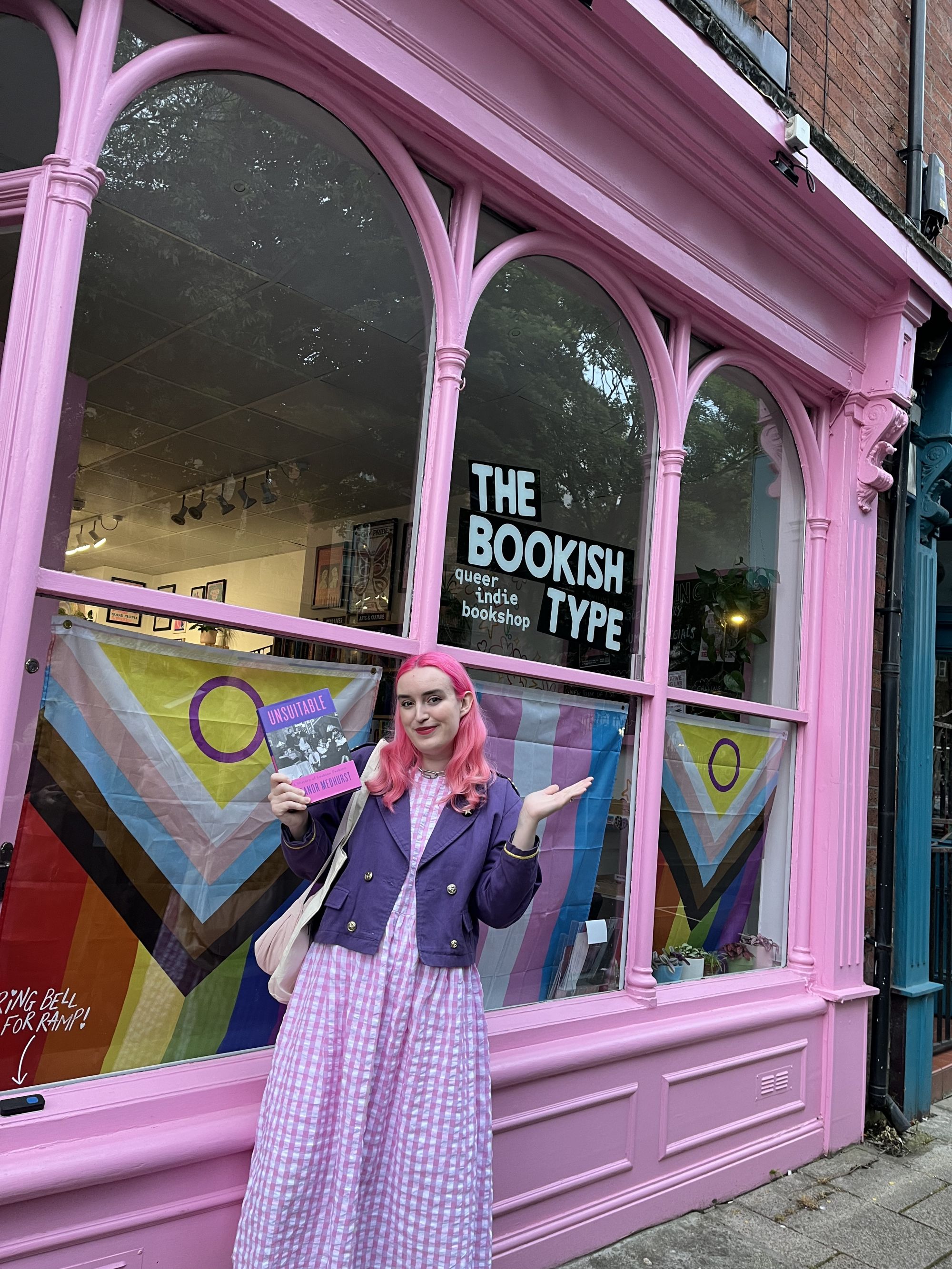 A woman with pink hair poses outside a pink bookshop holding a book