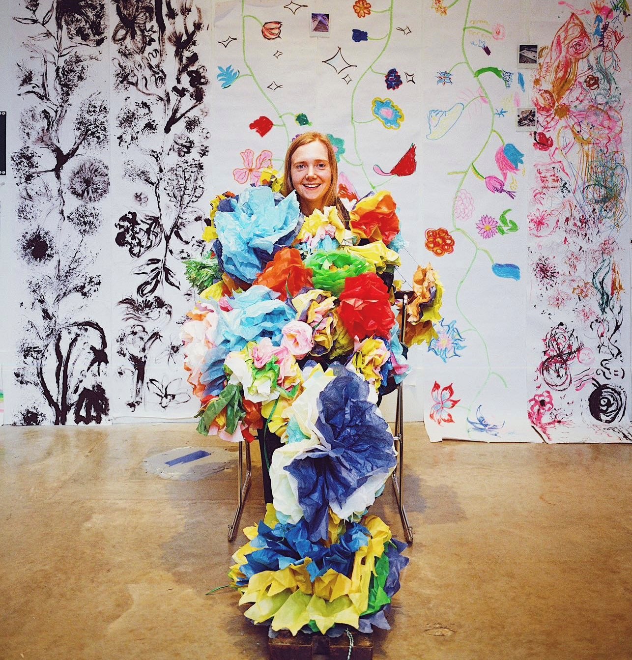A young blonde woman covered in colourful paper flowers smiles