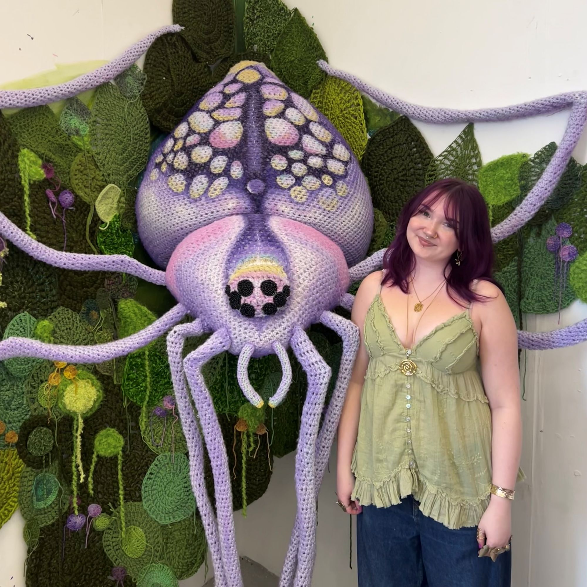 A young girl stands next to a giant purple knitted spider