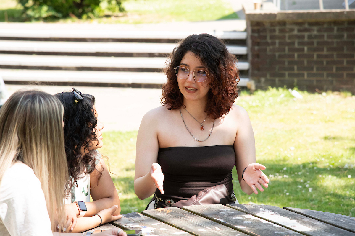 A young woman wearing glasses sits outside and speaks with her friends