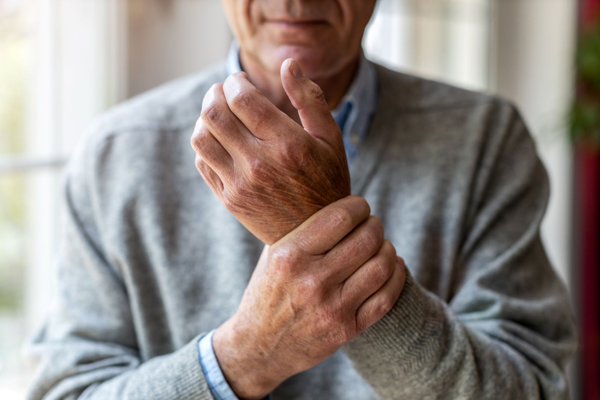 An elderly man wearing a jumper holds one wrist in his other hand. You can't see his face