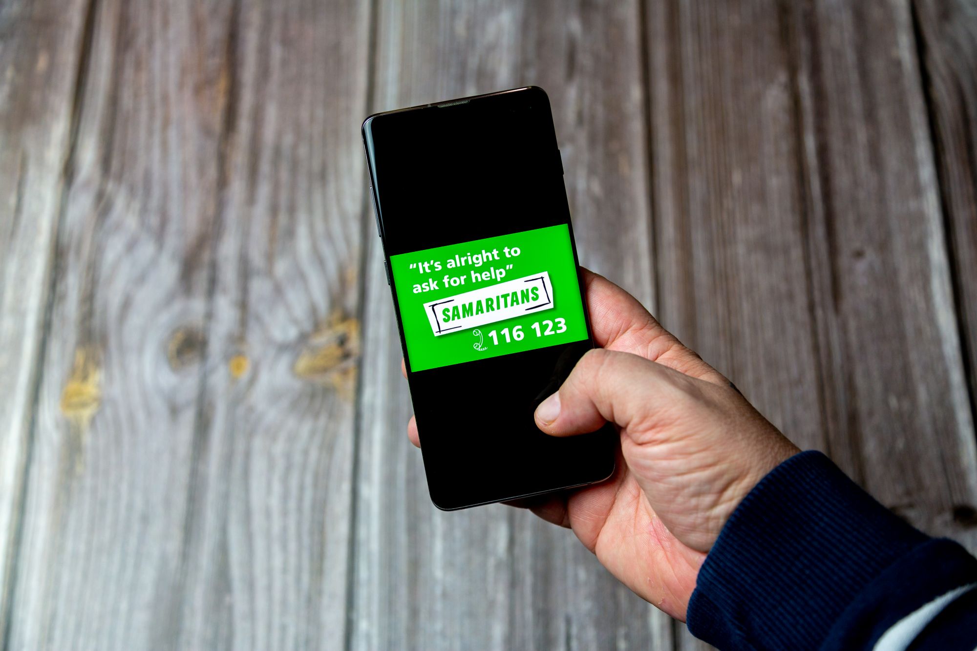 Hand holds a mobile phone which reads 'it's alright to ask for help - Samaritans'