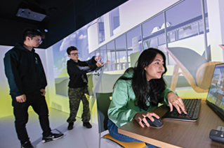 A group of students in a media space with projections all over the walls. One of the students is using a computer at a desk
