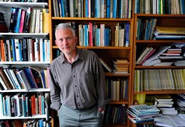 Man dressed in brown shirt and grey trousers standing in front of a bookcase
